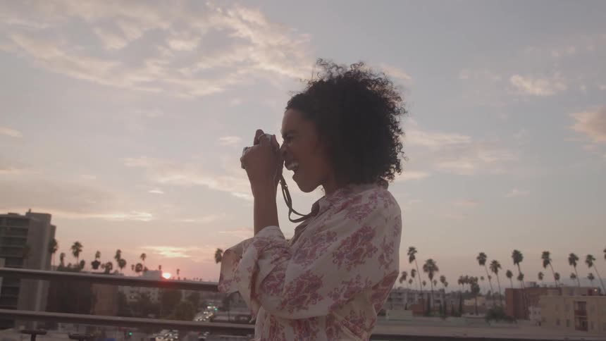 A woman on a rooftop takes a photo with a vintage camera during sunset