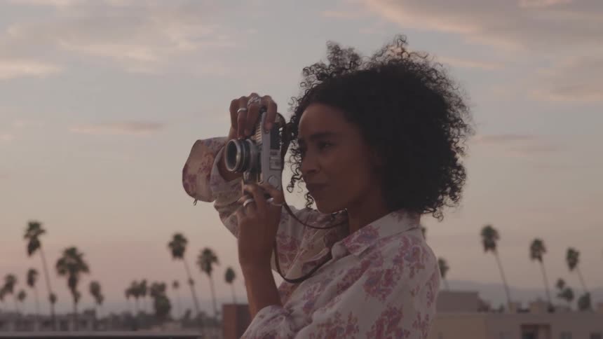 A woman with curly hair smiles as she holds a vintagelooking camera up to her face to take a picture she is standing on a rooftop at sunset with palm trees visible in the background