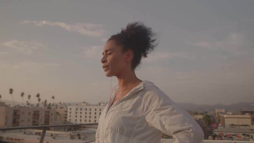 A young woman with curly hair and earbuds stands on a rooftop overlooking a city taking deep breaths and looking into the distance