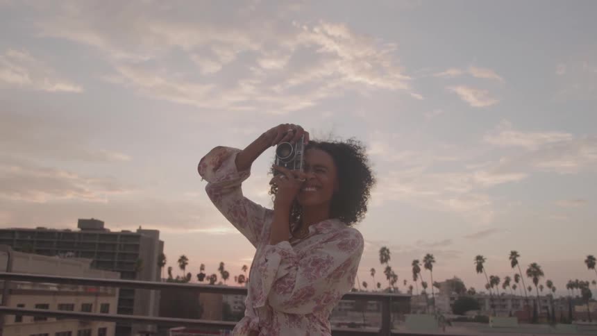 A woman on a rooftop holds a camera up to her face takes a photo and then lowers the camera while smiling