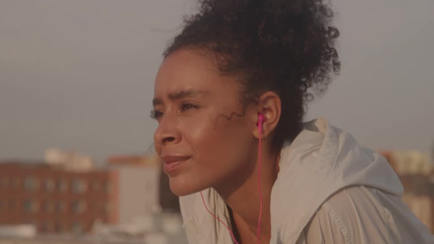Sweaty woman with a tired expression wearing pink earphones and a light jacket leaning forward after outdoor exercise