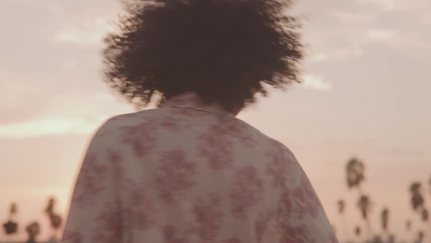 A woman with a large afro wears a patterned shirt and yellow headphones while she dances joyfully against a sunny sunset sky