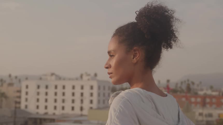 A woman is standing on a rooftop with her eyes closed and her face is looking toward the sky