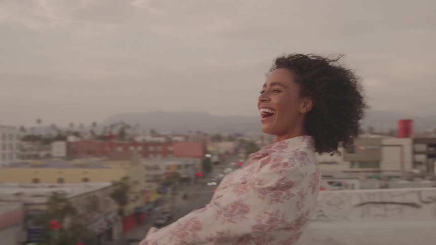A smiling person with curly hair is standing on a rooftop leaning against a railing theyre laughing and enjoying the view of the city below
