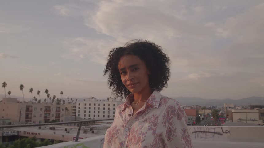 A woman stands on a rooftop at sunset overlooking the city