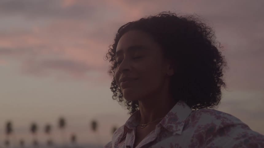 A young person with curly hair stands outside as the sun sets casting a warm glow they look pensive their eyes cast upward towards the sky