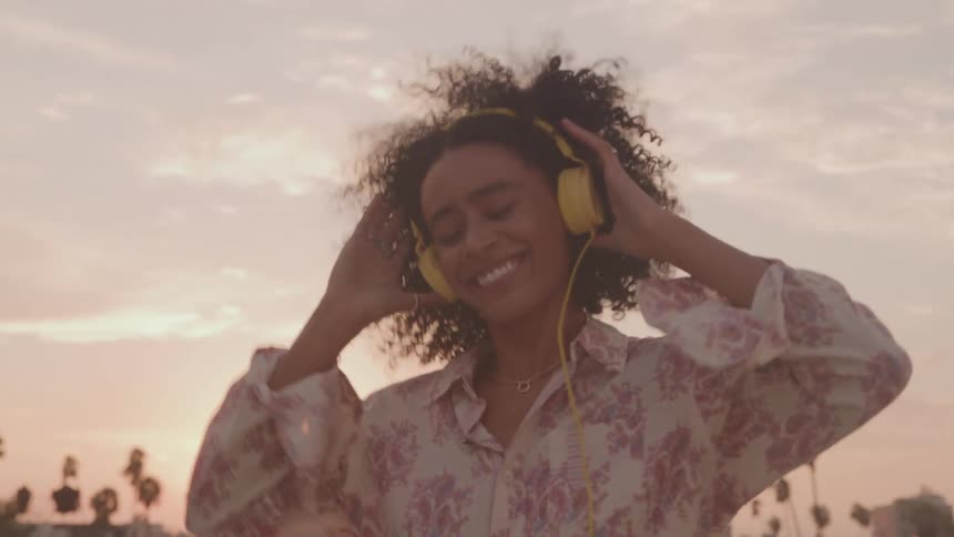 A young woman dancing in the middle of the street wearing a yellow headset lost in the rhythm as the orange setting sun casts a warm glow across the sky