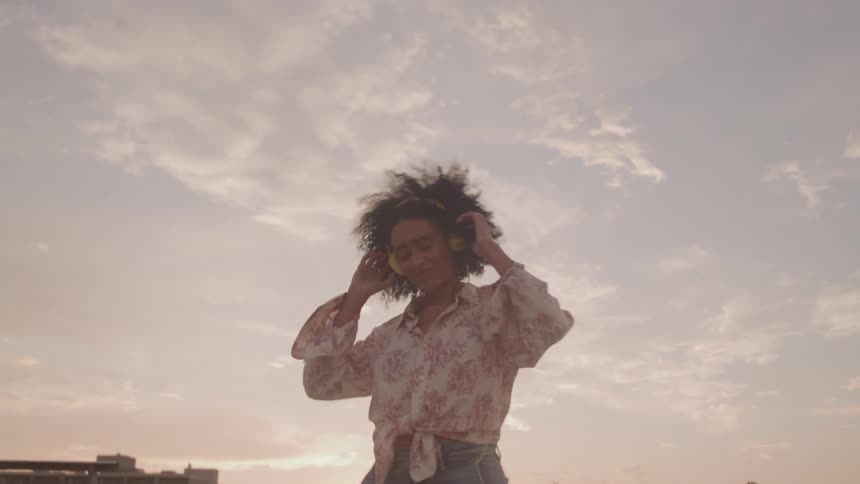 Close up of a young black woman dancing while listening to music with headphones on a rooftop at sunset