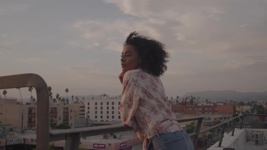 A woman leans on a rooftop railing gazing out over the city at sunset