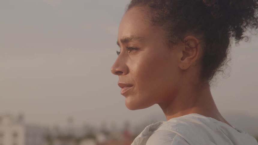 Woman in profile with tiedback hair gazes into distance under golden light with buildings and palm trees in background