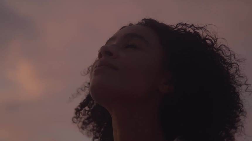 A person with dark curly hair looking up at the sky during sunset