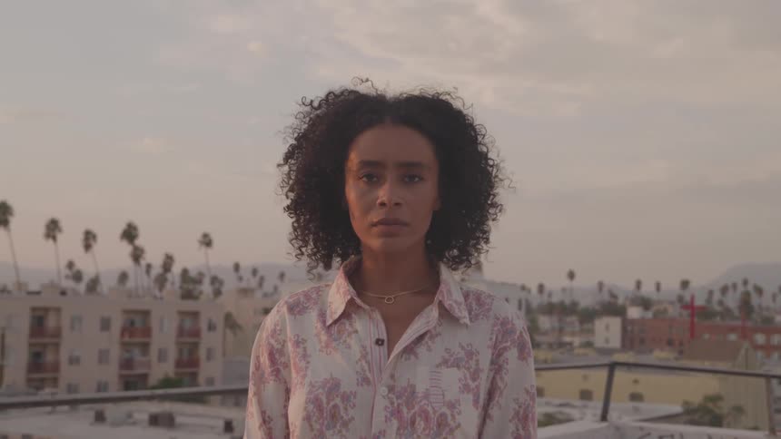 A young woman stands on a rooftop and looks at the camera with buildings and palm trees in the background