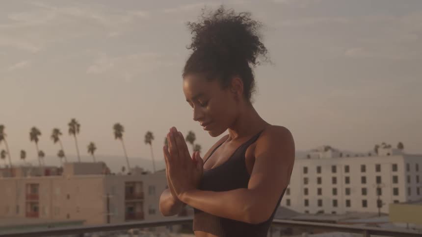 A medium shot shows a woman with her hair in a high bun practicing yoga on a rooftop she raises her arms into the air brings her hands together and then lowers them to her chest in a prayer position