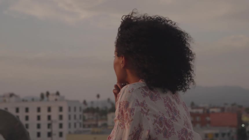 A medium shot shows a woman with curly hair on a rooftop she looks thoughtfully into the distance with the cityscape blurred behind her