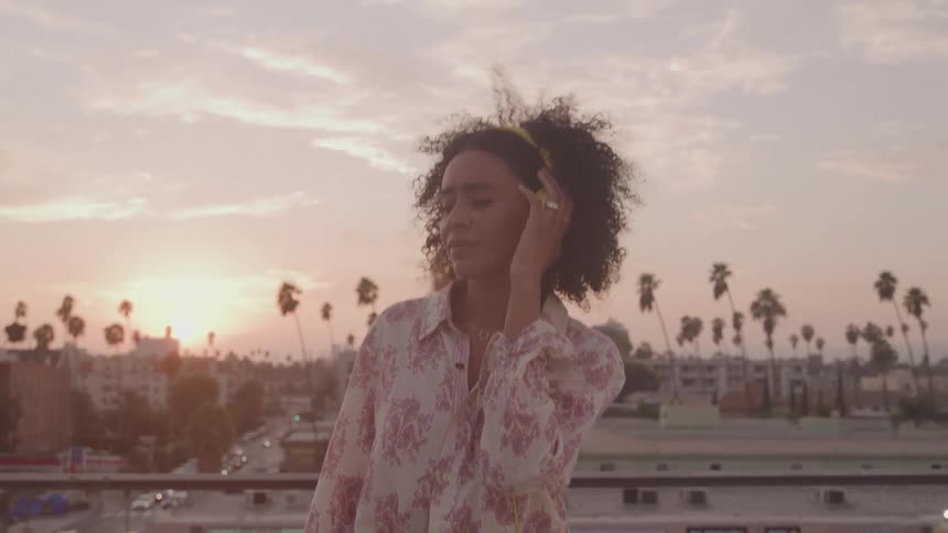 A person with curly hair is standing on a rooftop at sunset wearing yellow headphones theyre enjoying the music and the moment with a calm and peaceful expression
