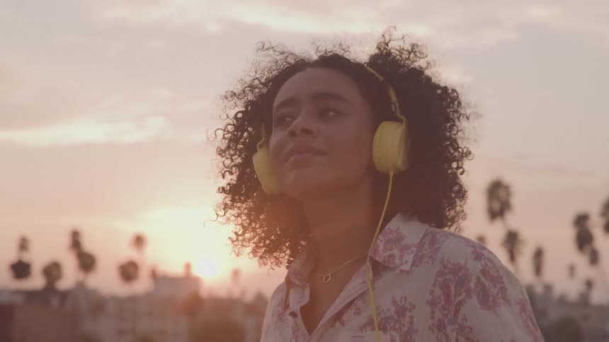 A woman enjoying music on a rooftop with yellow headphones at sunset