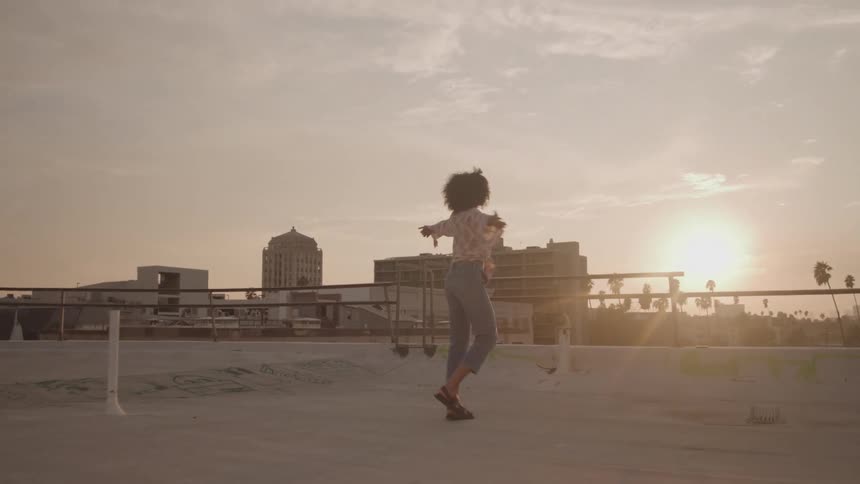 A woman with curly hair stands on a rooftop against the setting sun she moves her body expressively with her arms out wide taking in the moment of joy