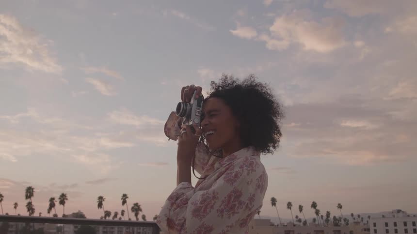 A woman is standing on a rooftop and holds a camera up to her face then lowers the camera