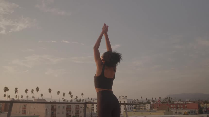 A young woman stands on a rooftop and stretches their arms in a yoga pose