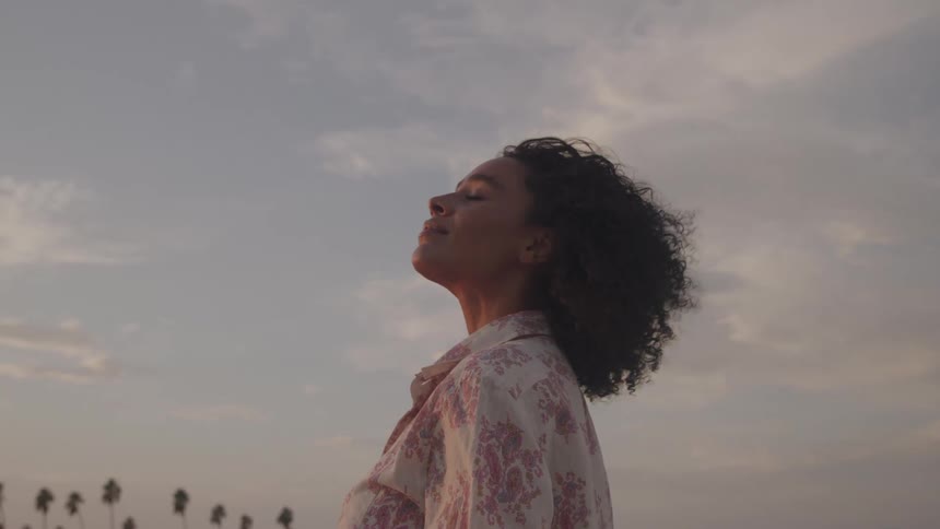 Young woman standing outdoors with long curly hair enjoying the warm air of nature at sunset