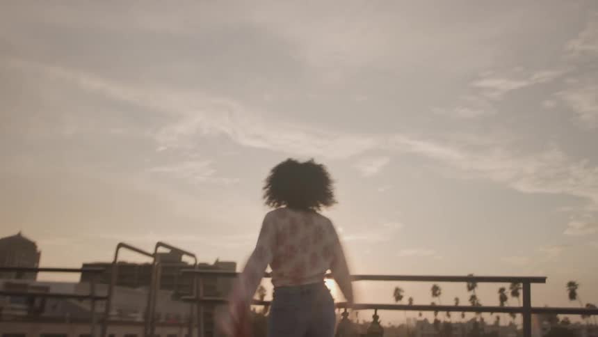 A full shot shows a woman with curly hair on a rooftop with a cityscape and palm trees visible in the background she raises her arms into the air and turns her head as if looking at the sky seemingly lost in a moment of pure joy