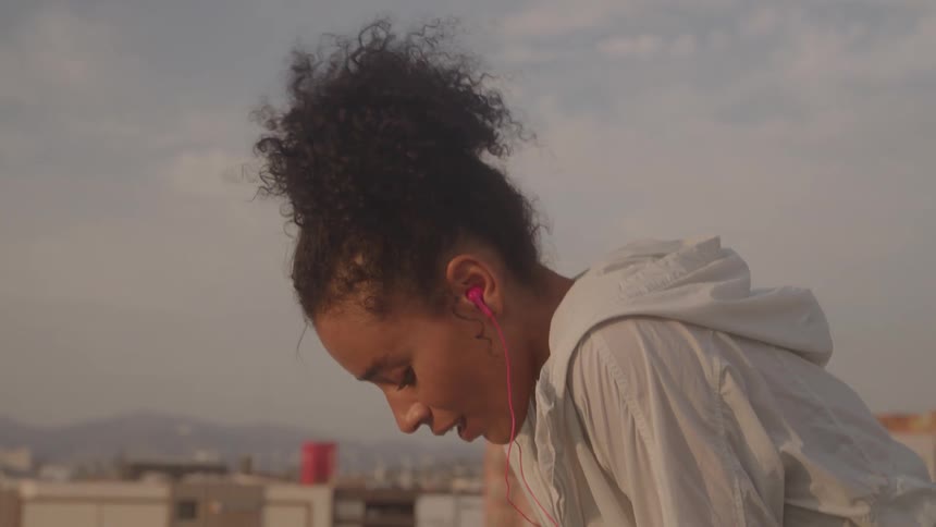 A woman with curly hair in a white hoodie and pink earbuds catches her breath on a rooftop after a workout