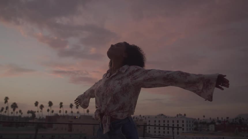 A young black woman spreading her arms in the air while standing outdoors on a rooftop at sunset