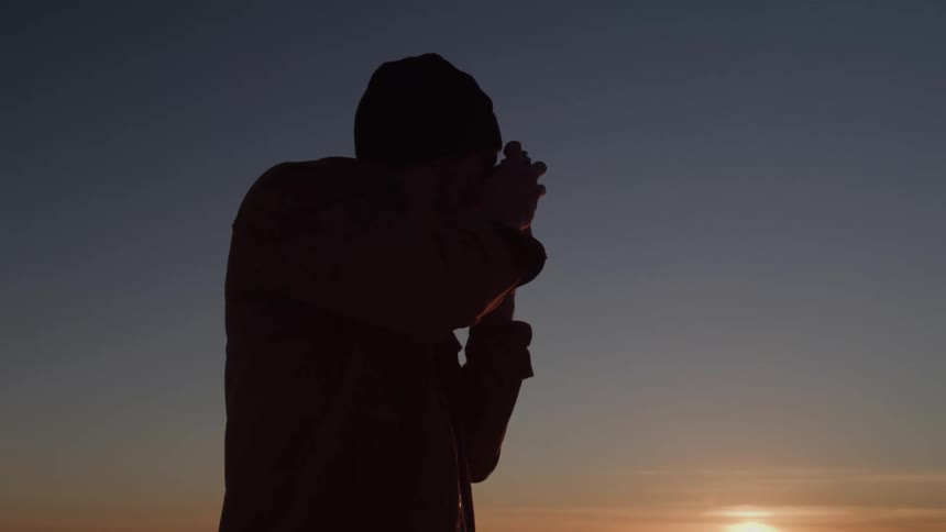 A silhouette of a person on a hill is holding a binoculars up to their face and looking in the direction of the setting sun