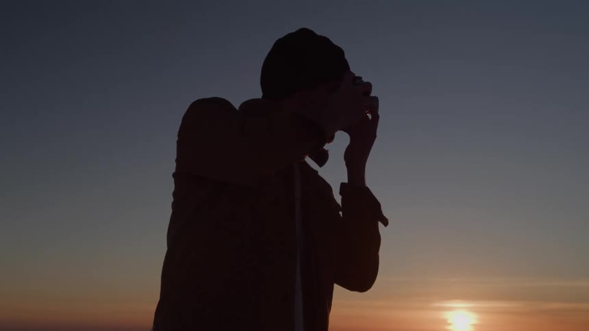 A silhouette of a person on a hill is holding a camera up to their face and looking in the direction of the setting sun
