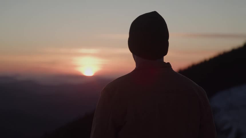 A man in a beanie watches the sinking sun standing still and reflective as if deep in thought