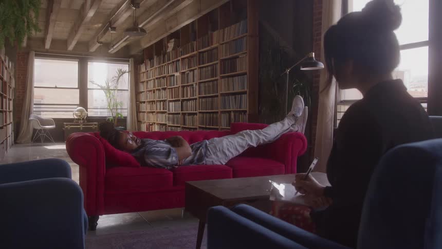 A person is lying on a red couch with their feet up while talking to a person who is sitting in a chair and writing on a notebook