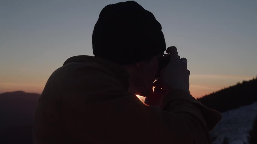 Close up of a young man taking photos with a camera while standing outdoors at sunset