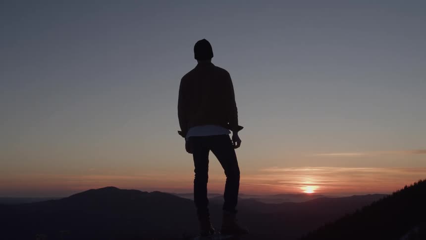 A person stands silhouetted against a mountain sunset looking out at the horizon the vibrant sky fades from a warm orange at the horizon to a cool blue overhead with jagged mountains visible in the distance