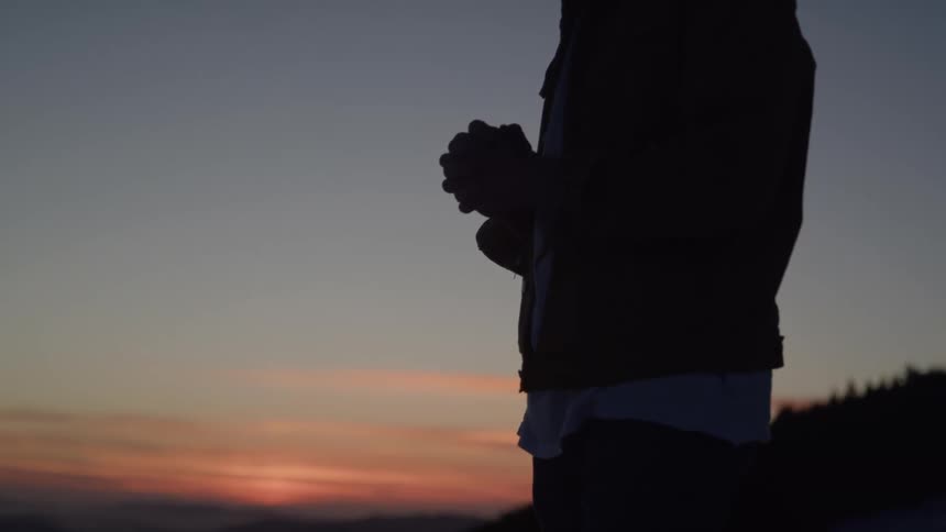A man clasps his hands to his chest silhouetted against a soft sunset his posture quiet and pensive