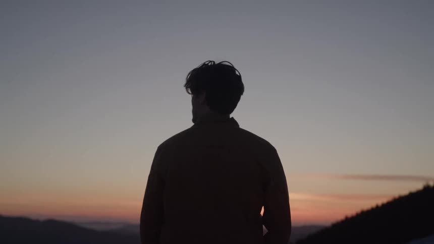 A man with his back to the camera standing and looking out over a mountain range at a vibrant sunset