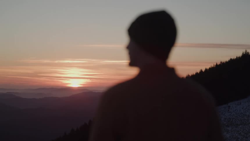 A dark video shows the blurry silhouette of a man wearing a beanie looking out over a mountain range at a vibrant sunset