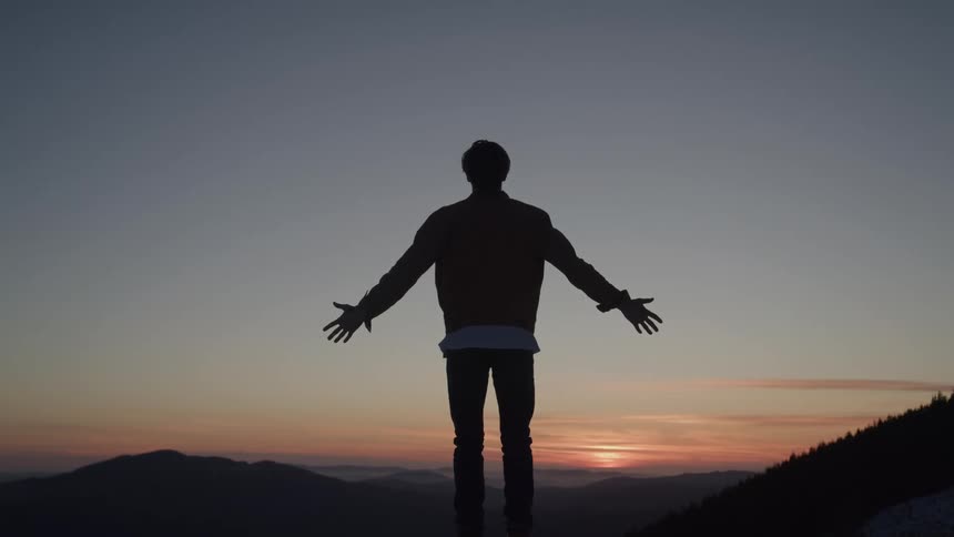 A man stands silhouetted on a ridge at sunset with his arms outstretched looking serene and quietly triumphant