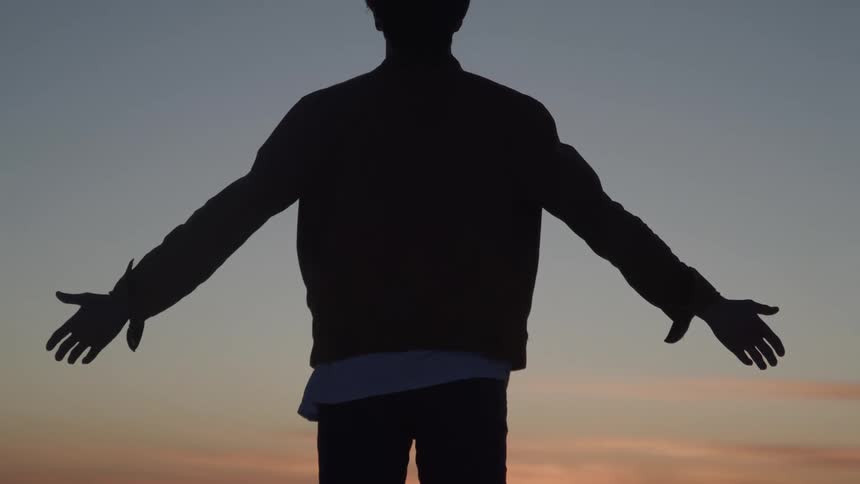 A silhouette of a person standing outside with their back to the camera and their arms are out to their sides