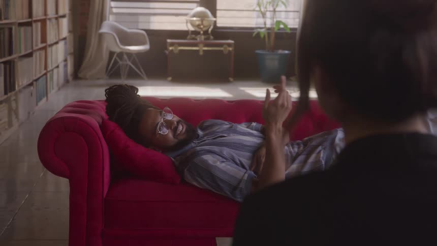A full shot captures a man with dreadlocks lying on a red couch gesturing with his hands as he speaks to someone the room is filled with light and lined with bookshelves