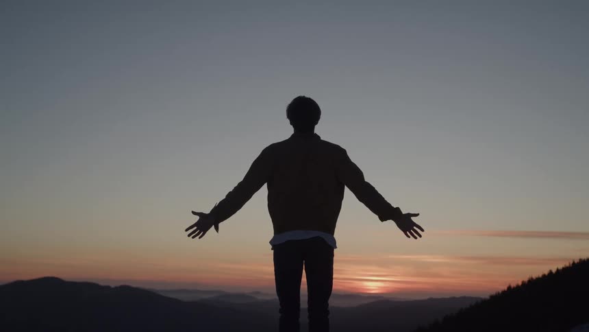 A silhouette of a man with his back to the camera stands on a mountain at sunset and raises his arms as if welcoming the view of the valley below