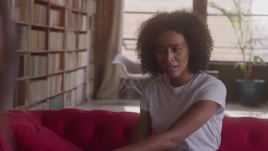 A woman with curly hair on a red couch expresses frustration while talking to someone off camera in a sunlit room with bookshelves
