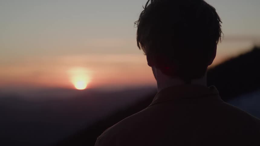 A man stands on a ridge silhouetted against the sunset looking thoughtful and quietly resolute as he surveys the distant hills