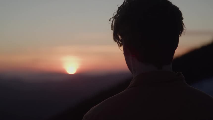 A silhouette of a person is shown from behind as they watch a sunset the sun is a bright orb casting a warm orange glow across the sky as it descends behind a mountain range