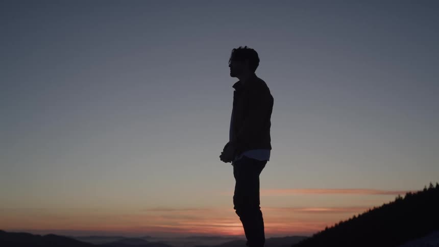 A man stands silhouetted on a ridge at sunset hands loosely clasped and shoulders slack looking quietly contemplative