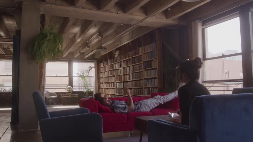 A full shot captures a man with dreadlocks lying on a red couch gesturing with his hands as he speaks to someone the room is filled with light and lined with bookshelves