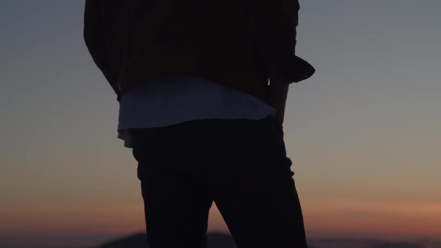 A silhouette of a person standing outside at sunset is holding a hand in a pocket