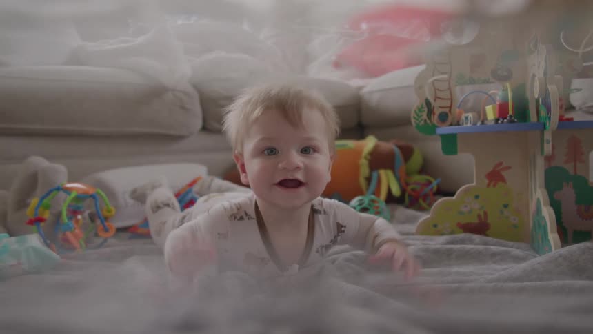 A cute baby with bright blue eyes crawls playfully on a blanket surrounded by toys in a bright living room