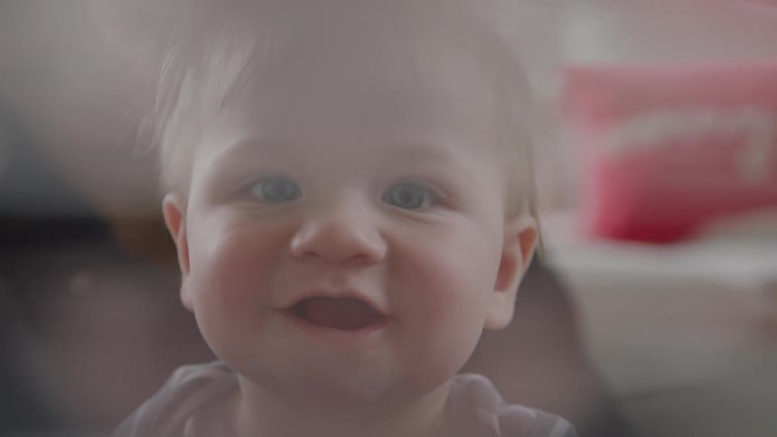 A smiling baby with light hair and blue eyes looks directly at the camera