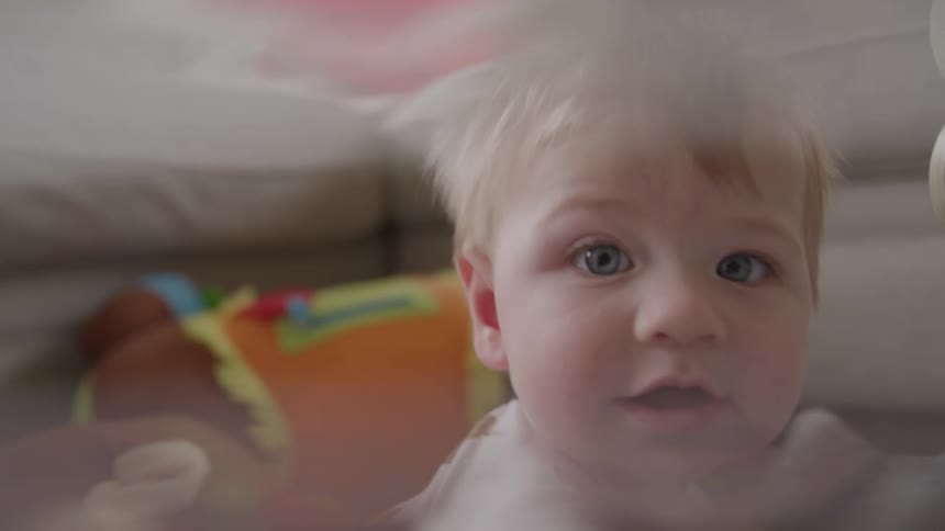 A happy baby with blue eyes and light hair smiles at the camera then gets excited and moves closer