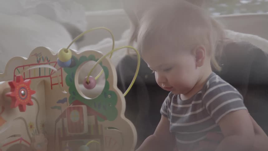 A baby plays with a colorful wooden activity toy while being held by an adult in a soft bright setting